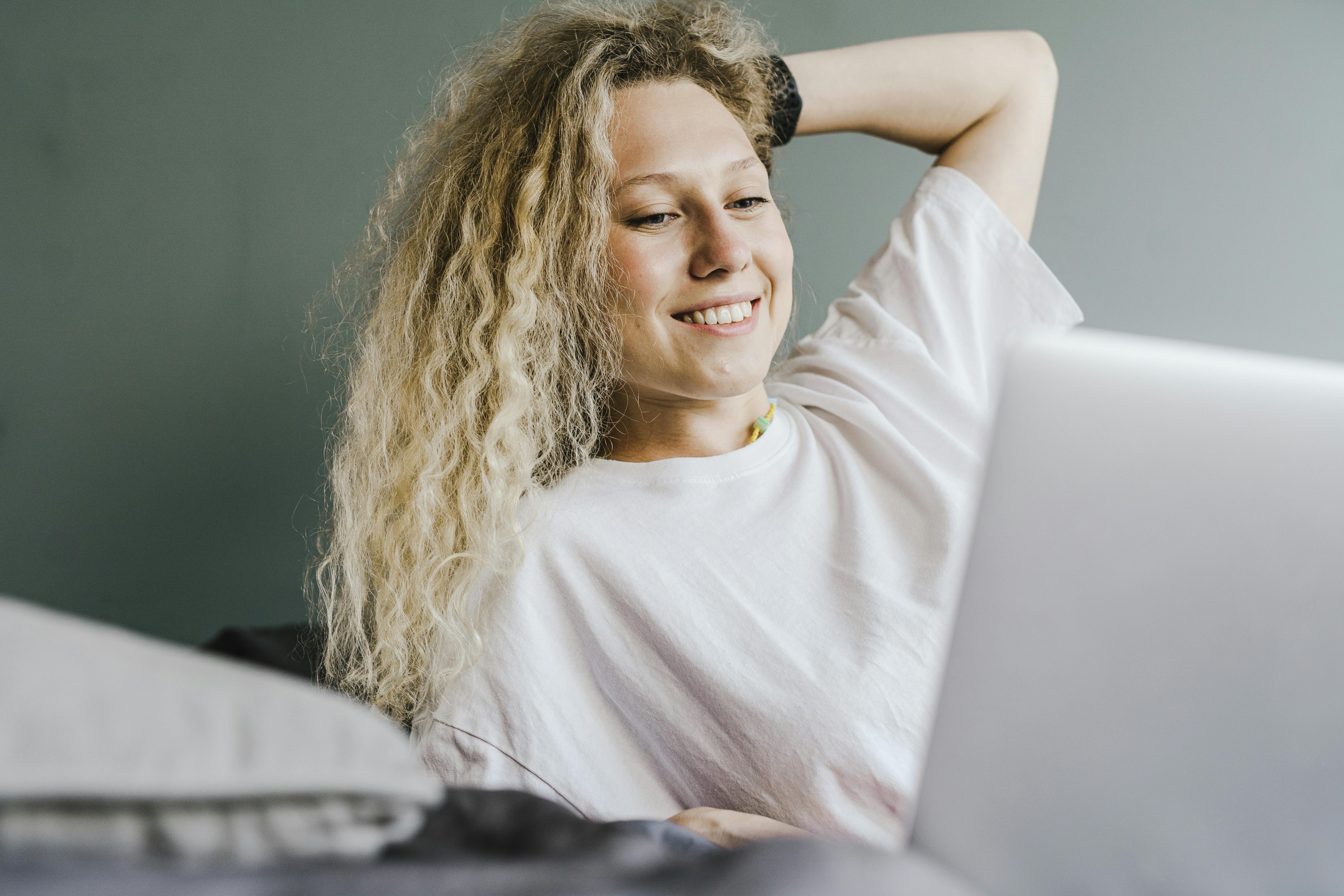 A cheerful woman with curly hair lounges comfortably, enjoying her leisure time on a laptop indoors.