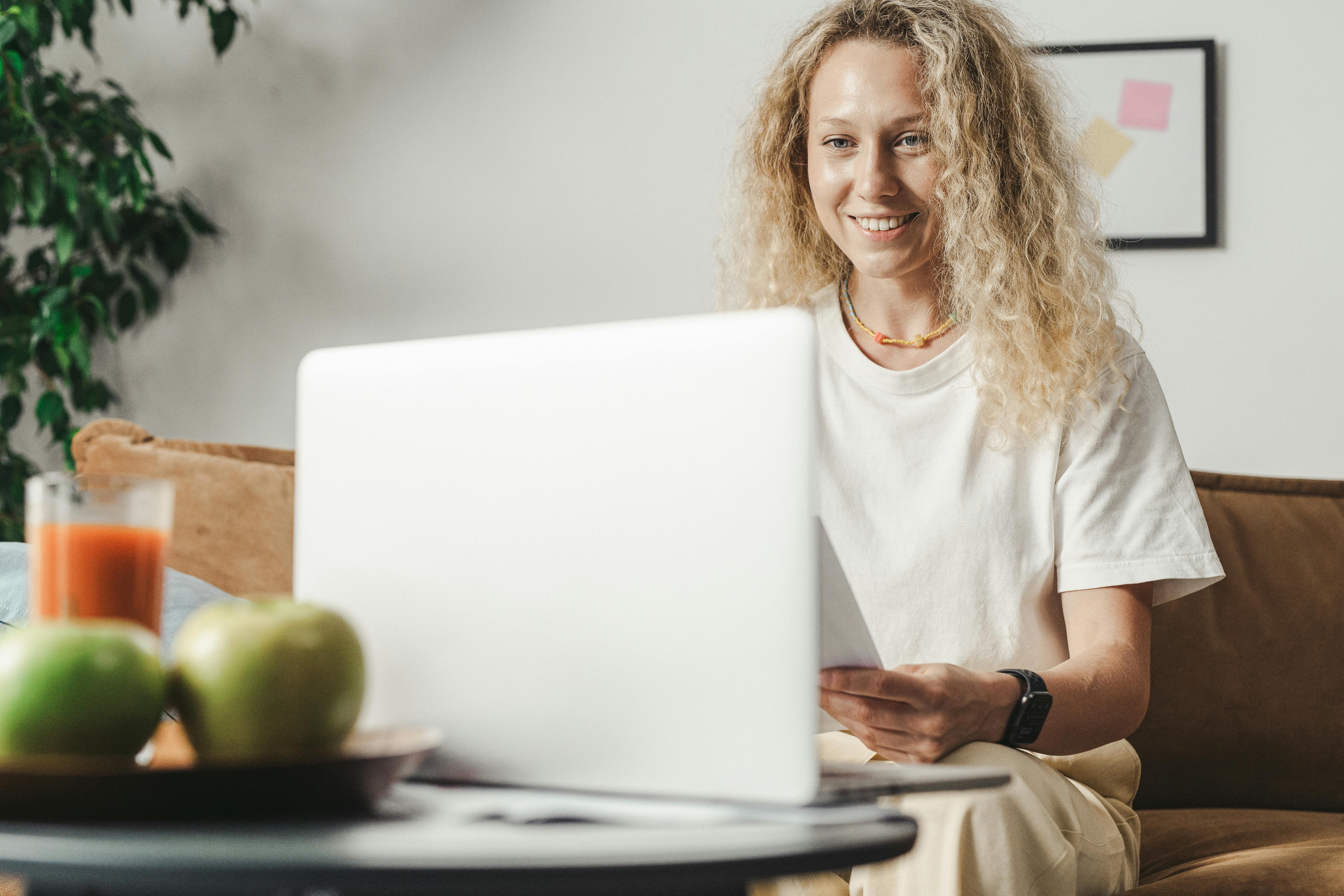 A woman with curly hair smiling as she works on her laptop indoors, complementing a relaxed freelance lifestyle.