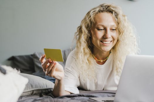 Smiling blonde woman with curly hair shopping online with a bank card indoors.