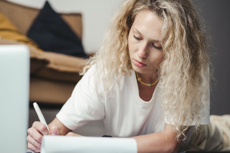 Young woman with curly blonde hair writing at a desk indoors, focused on her task.