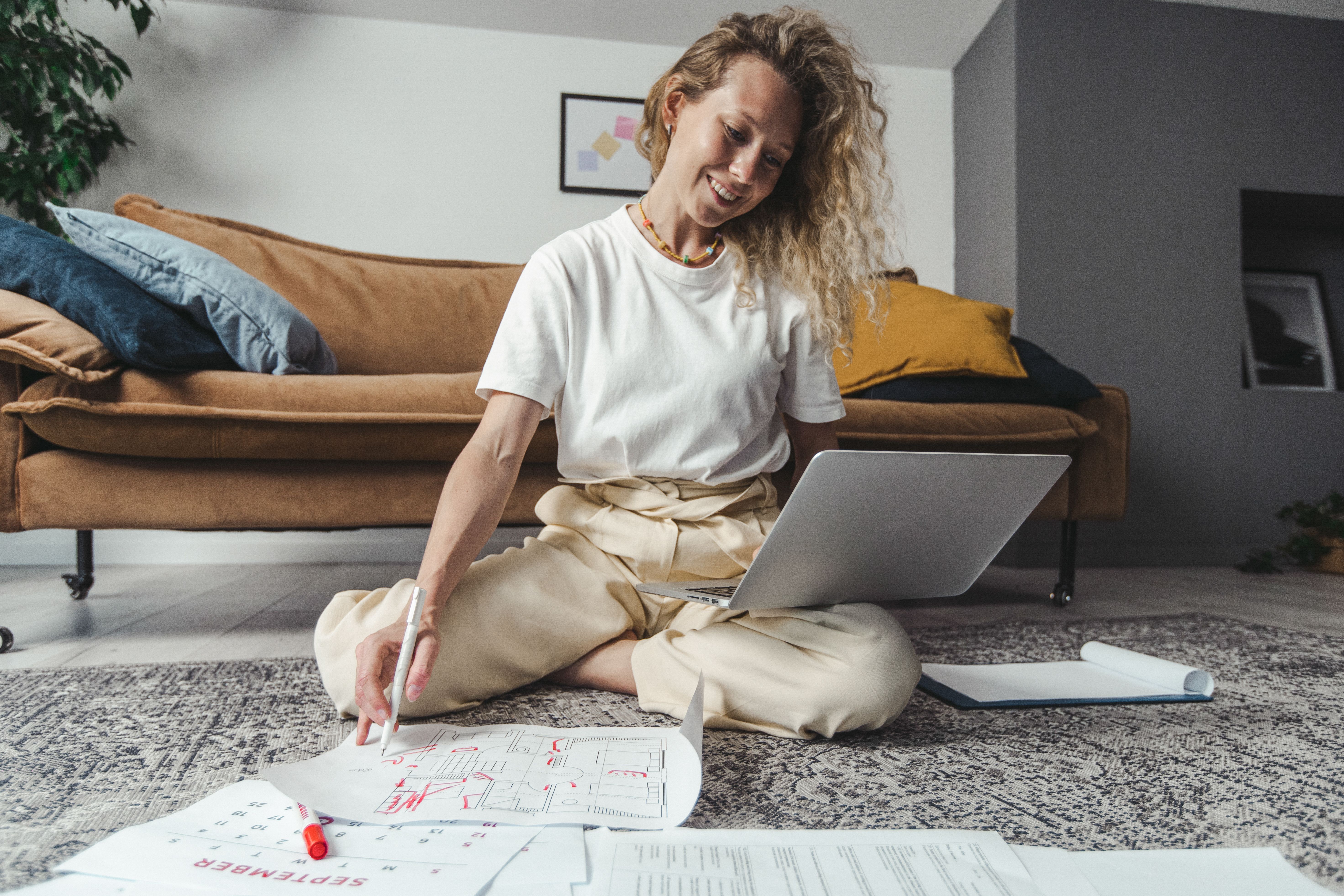 Smiling woman sitting on floor, working on laptop, surrounded by papers in cozy home setting.