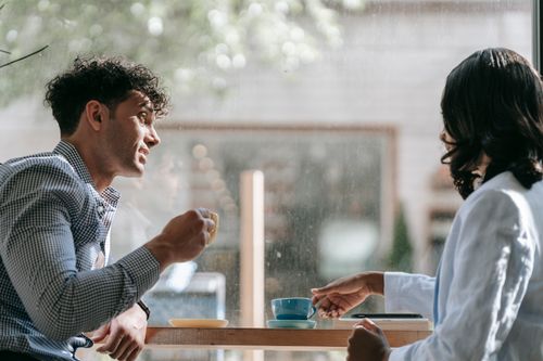 Free Two adults enjoying a casual conversation over coffee in a cozy café with natural lighting. Stock Photo