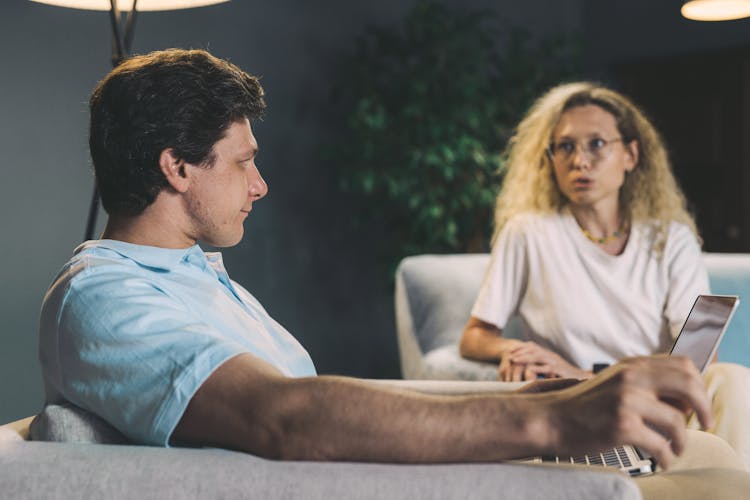 Man Sitting On A Sofa Holding A Laptop And Woman Sitting Near And Talking To The Man 