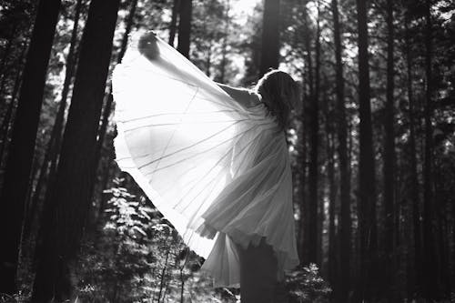 Grayscale Photo of Woman in White Dress Standing in Forest