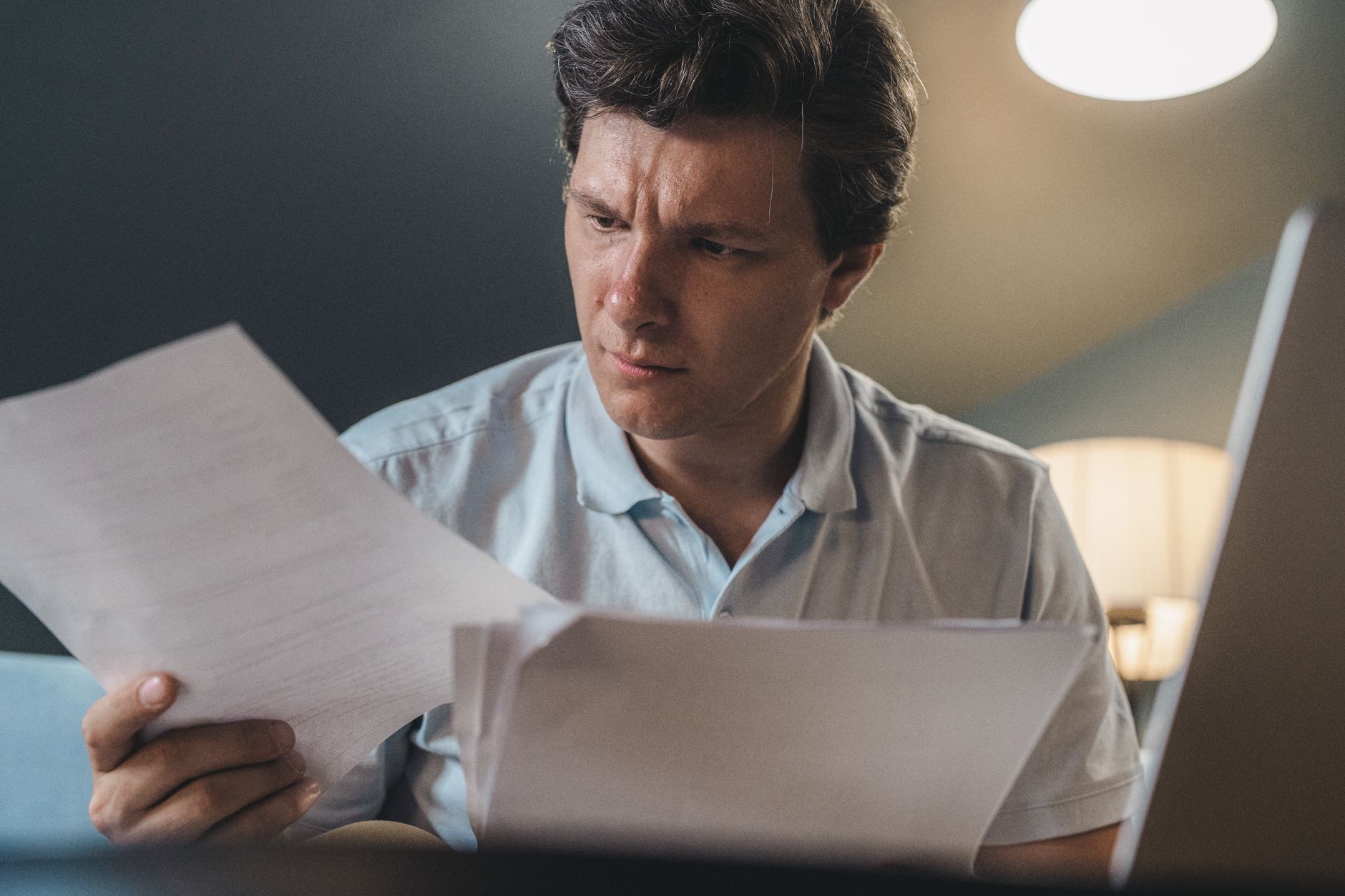 A man intently examines papers, seated indoors under warm lighting, focusing on his work.