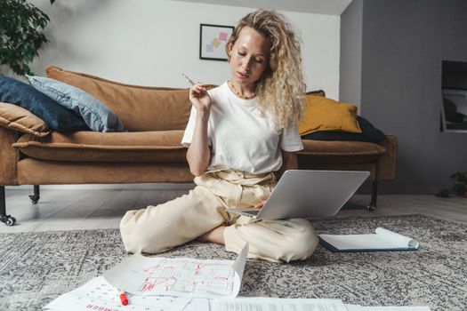 A woman working on a laptop while sitting on the floor with papers around, in a cozy home setting.