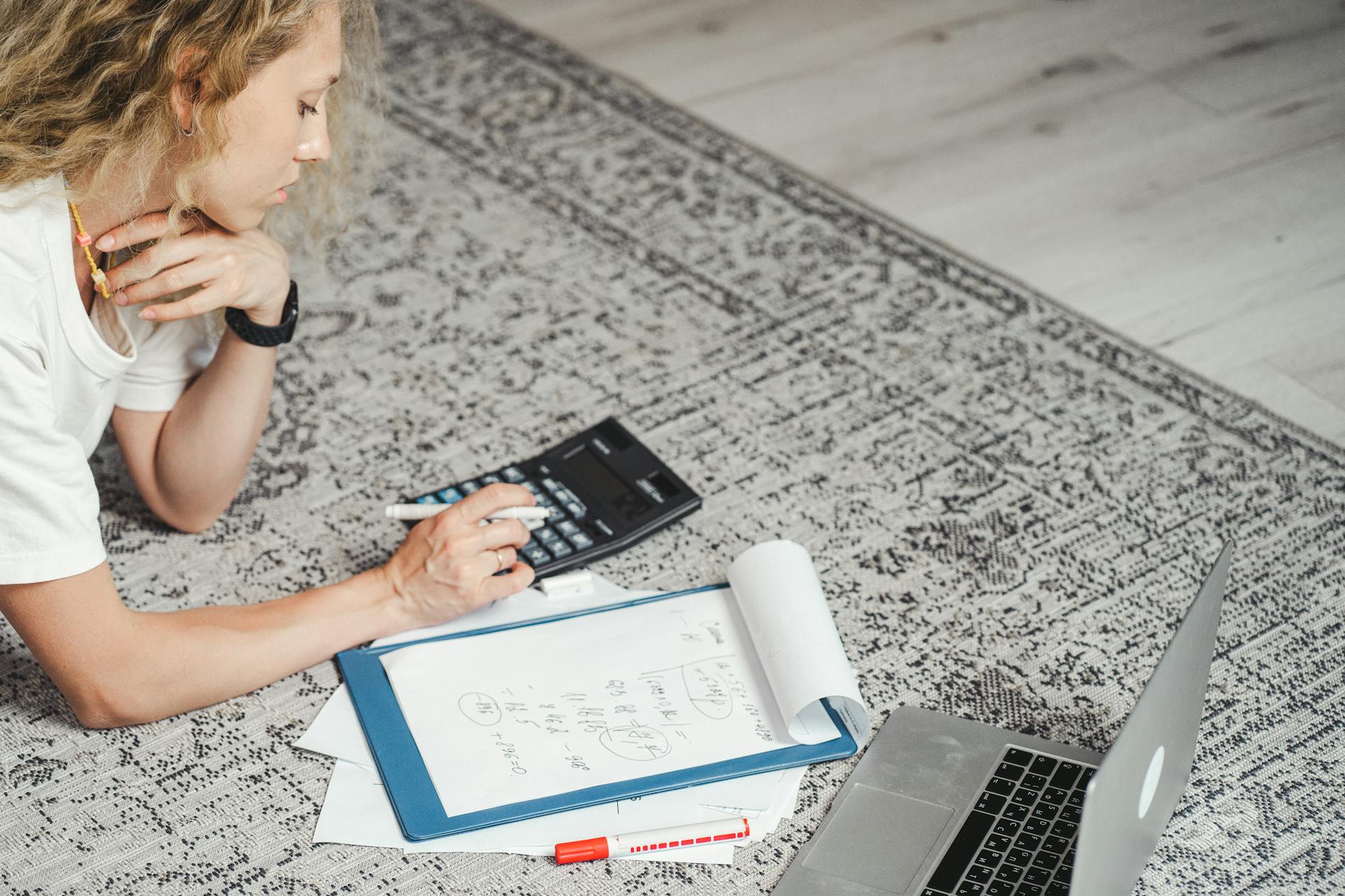 A woman calculates finances at home using a laptop, calculator, and documents on a rug.