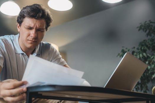 Man focusing intently while reviewing documents at a desk with a laptop.