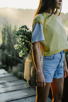 Stylish young woman wearing casual summer outfit with a net bag, standing on a wooden path in nature.