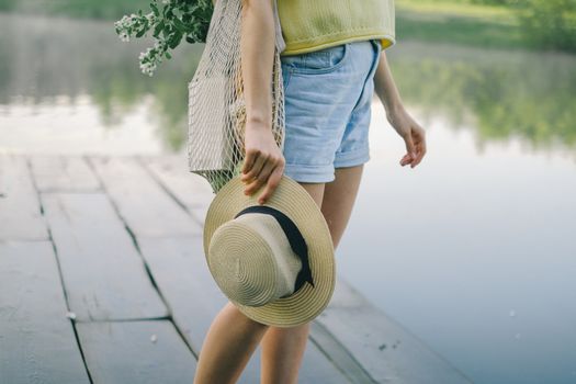 A woman standing by the lake in denim shorts, holding a straw hat and a net bag.