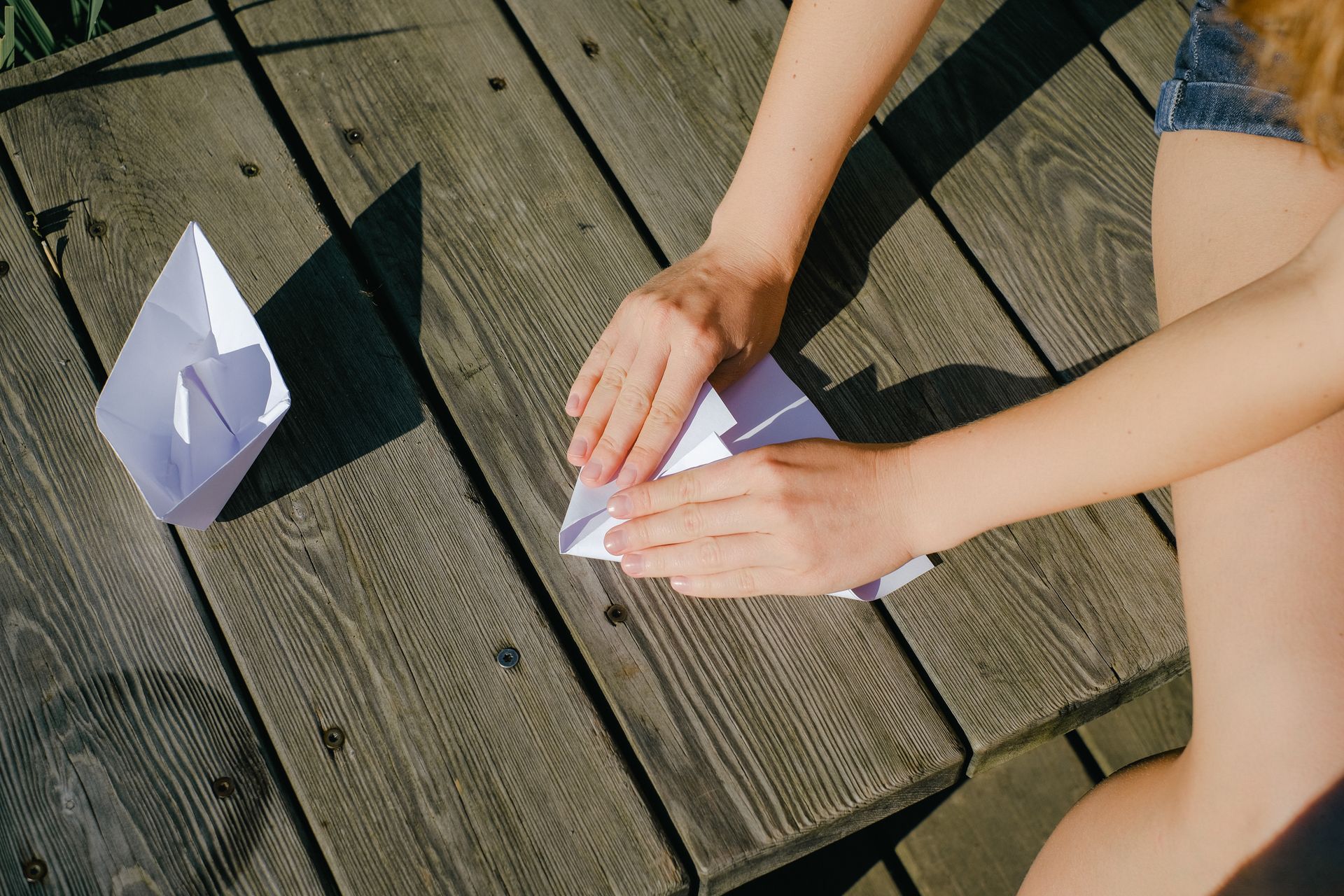 Hands folding paper boats on a wooden deck under the sun. Creative and peaceful outdoor activity.
