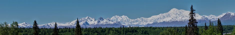 Breathtaking panorama of snow-capped mountains and forest under clear blue sky.
