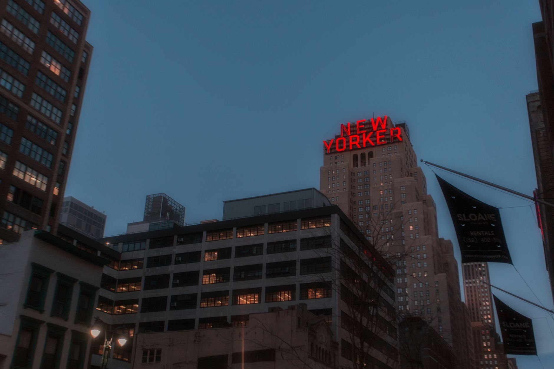 Capture of the New Yorker Hotel neon sign amidst skyscrapers at dusk in New York City.