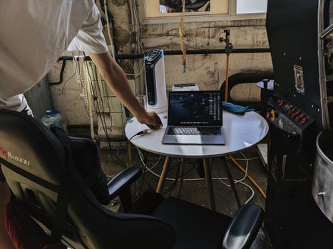 A workspace with a laptop, electronics, and cables in an industrial room with dim lighting. One person visible.