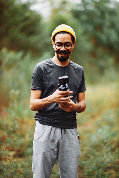 Free Bearded man in yellow beanie holding a camera, smiling outdoors. Stock Photo