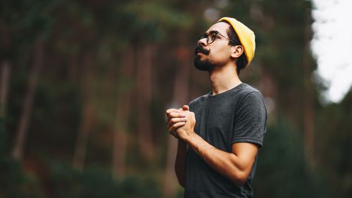 Free A man looks up thoughtfully in a forest, wearing a yellow beanie and glasses. Stock Photo