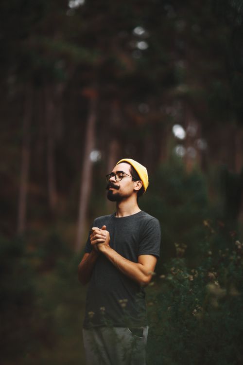 Free A man with facial hair and a yellow beanie stands in a forest, hands together, eyes closed. Stock Photo