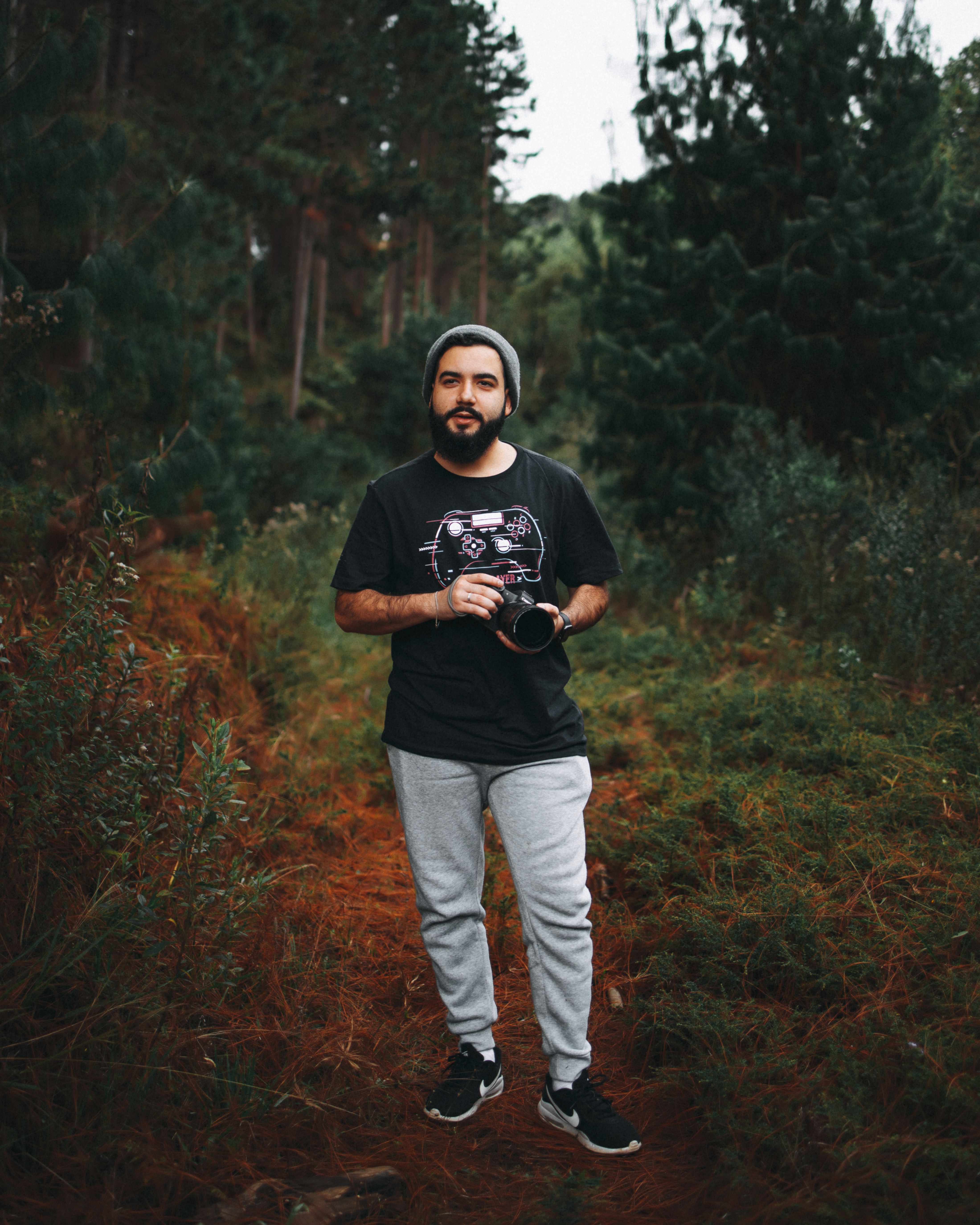 A bearded man holding a camera, standing in a lush forest, exuding a contemplative mood.