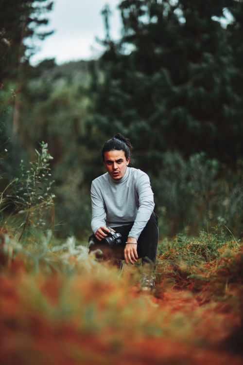 Free A man squats in a lush forest while holding a camera, surrounded by greenery. Stock Photo