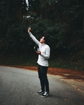 A man controlling a drone on a rural roadside, surrounded by lush trees.