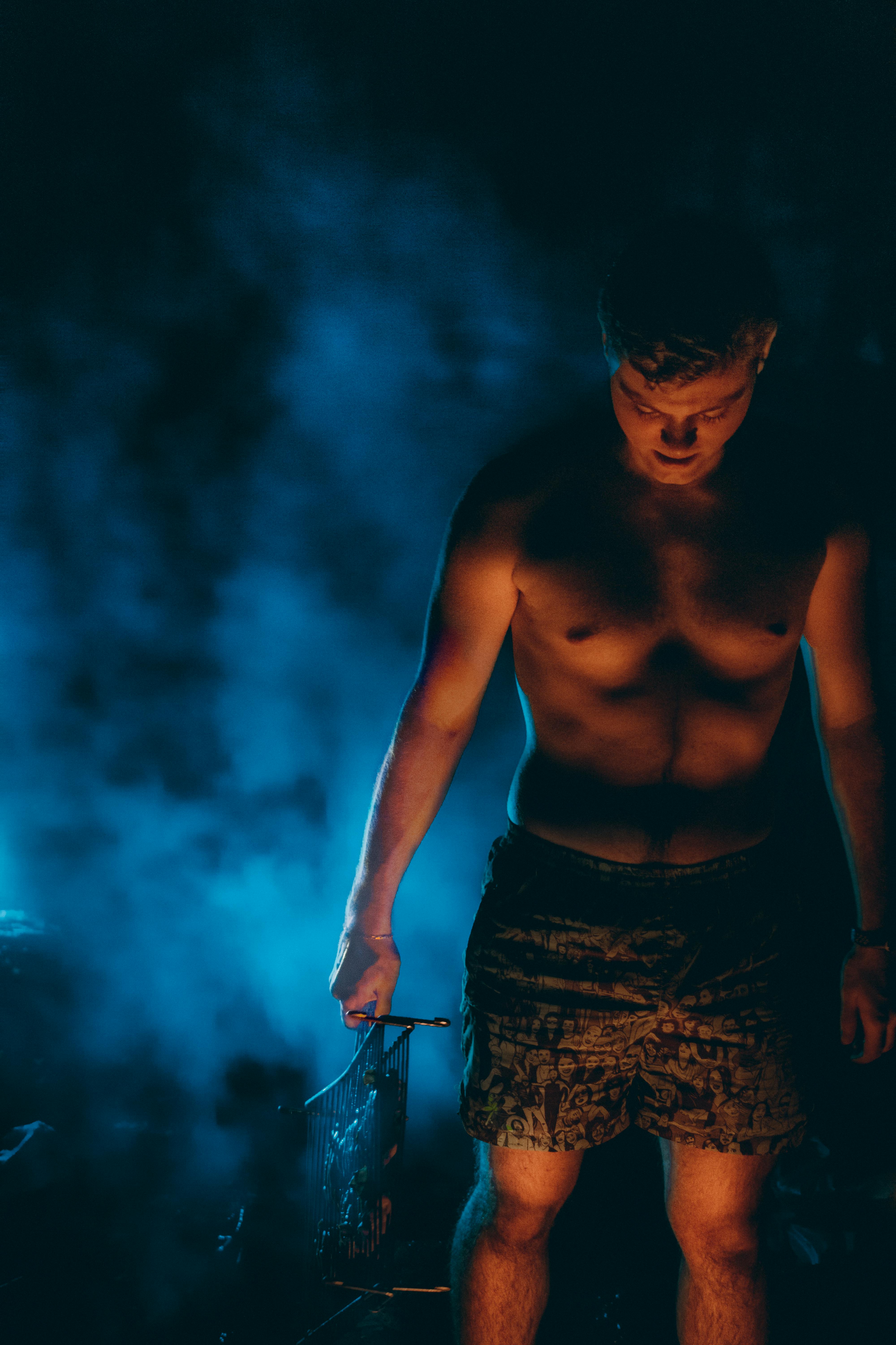 A shirtless man stands surrounded by blue smoke, creating a dramatic and moody scene.