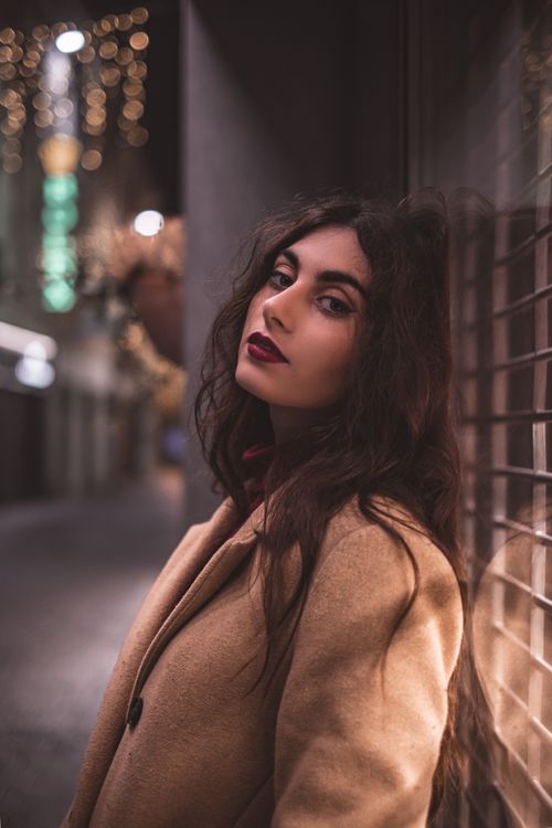 Free Stylish brunette woman leaning against a glass window at night in an urban setting. Stock Photo
