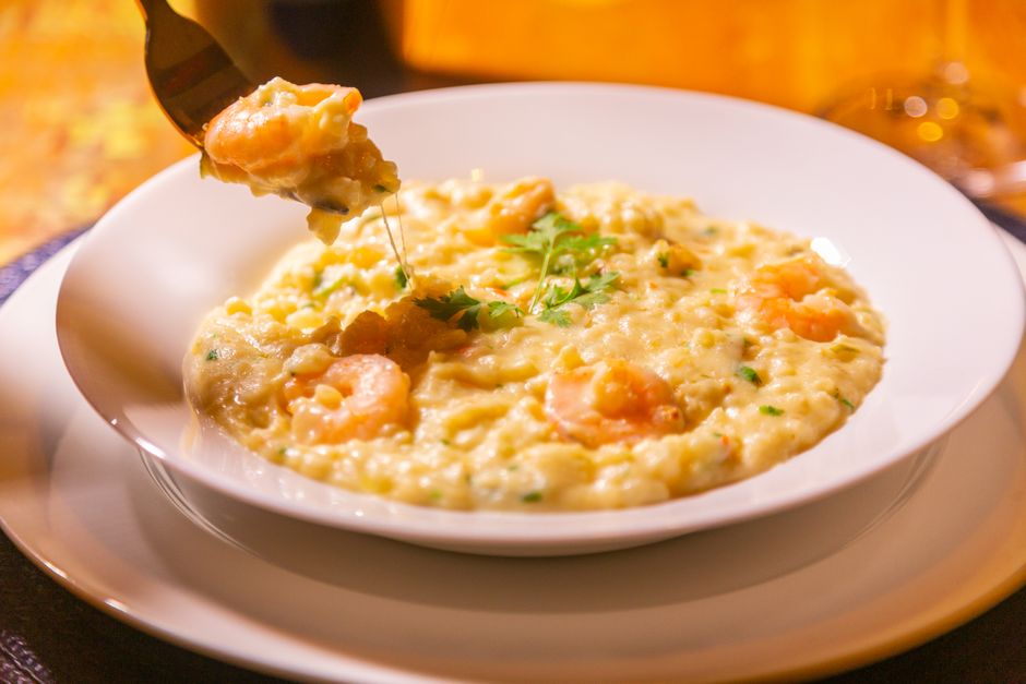A white bowl of creamy pale-yellow cheese grits topped with a pat of melting butter and a crack of black pepper, steam rising, set on a wooden table.