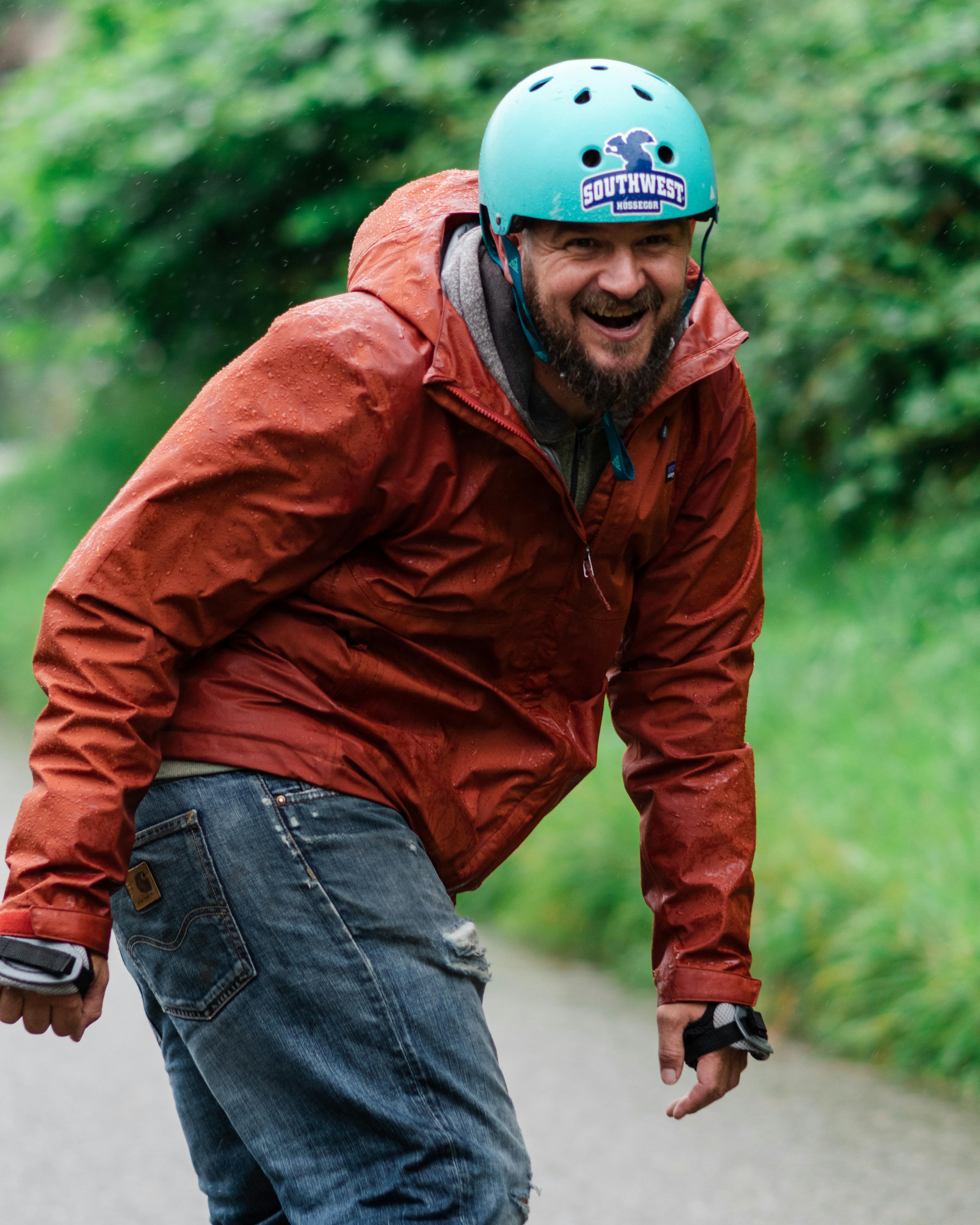 Bearded man enjoying skateboarding outdoors in an orange jacket and helmet, embracing adventure and joy.