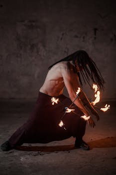 Topless man performs with flaming torches in a dark room, capturing an intense fire dance moment.