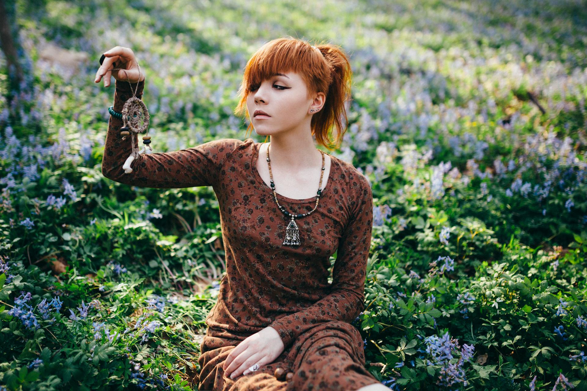 Woman in Brown Long Sleeves Dress Sitting on Green Grass Field while Holding a Dream Catcher