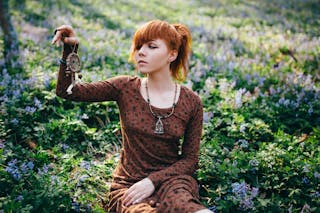 Woman in Brown Long Sleeves Dress Sitting on Green Grass Field while Holding a Dream Catcher