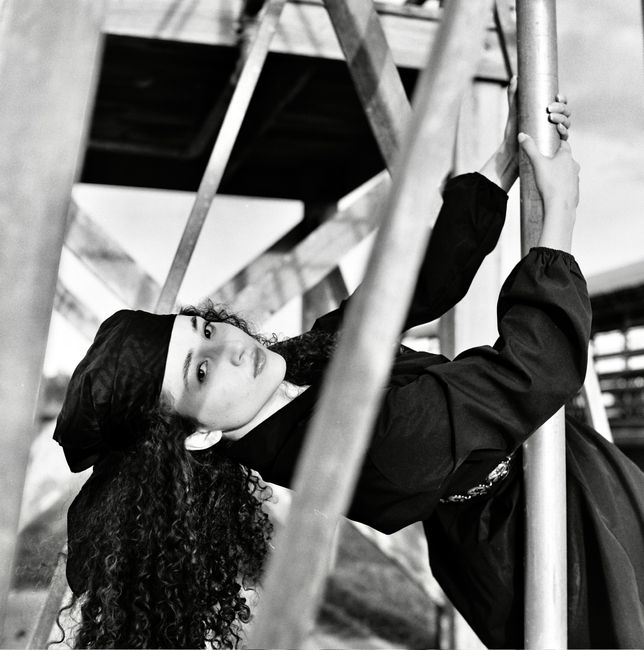 Striking monochrome portrait of a graduate leaning against a metal framework, capturing a blend of elegance and industrial aesthetics.