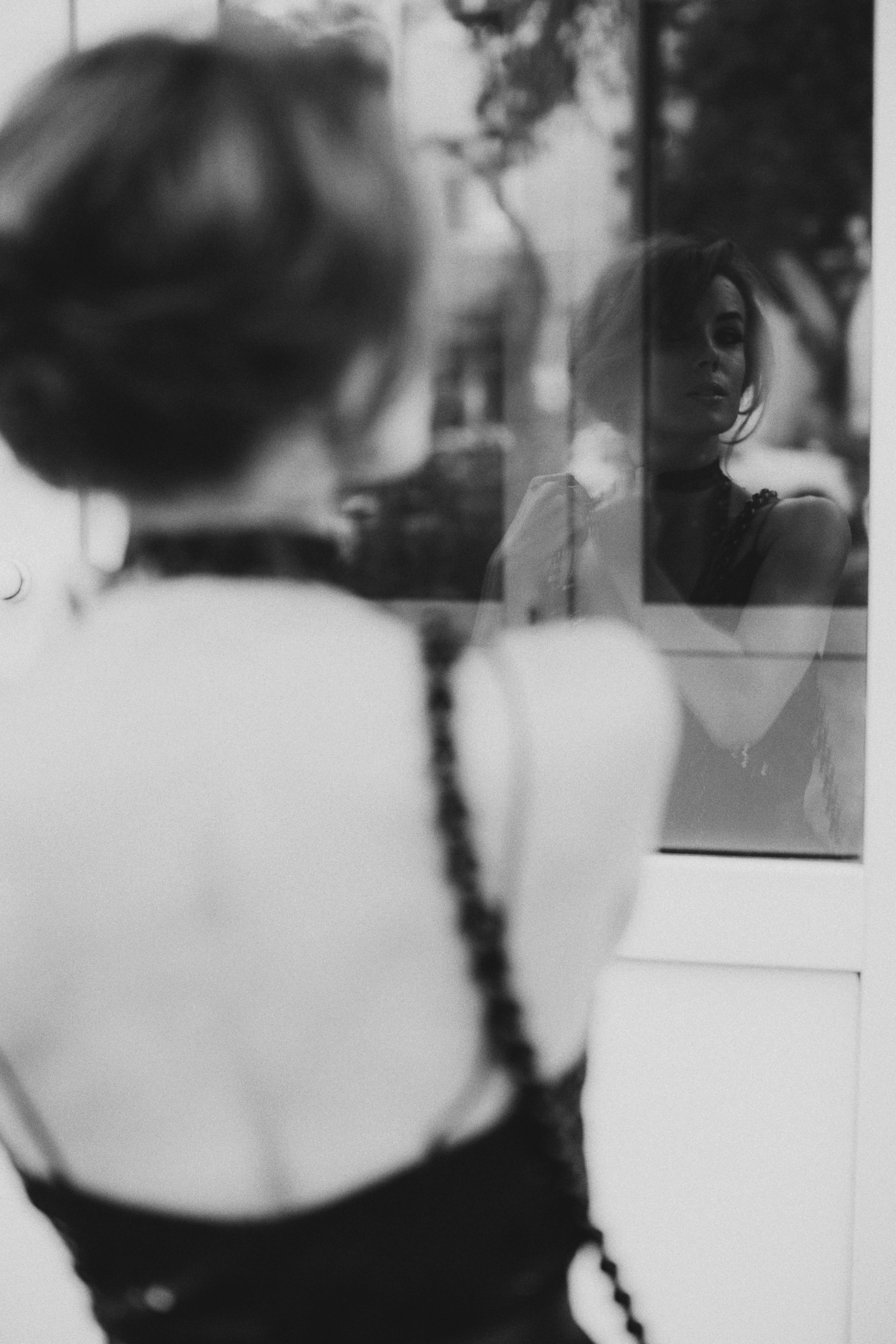 A stylish woman in a backless dress is elegantly reflected in glass, captured in grayscale.