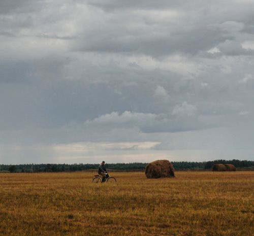 Free A lone cyclist rides through a tranquil countryside field under a cloudy sky with haystacks. Stock Photo