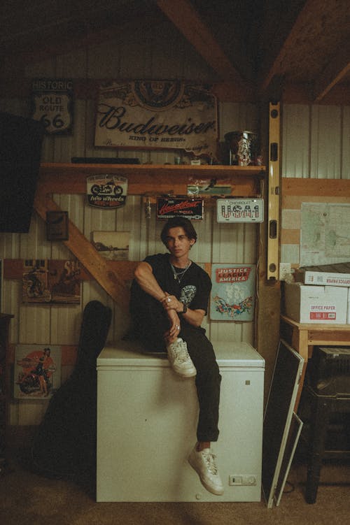 Man in Black Crew Neck T-shirt and Pants Sitting on Chest Freezer with Vintage Collections on Wall