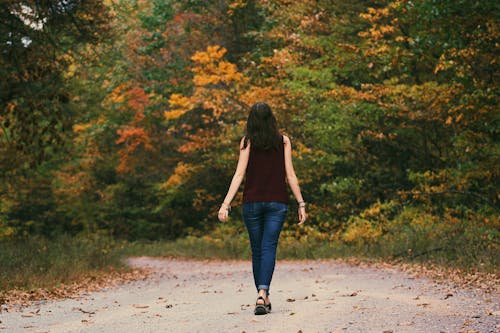 Женщина в коричневом платье без рукавов и синих джинсах стоит на серой дороге