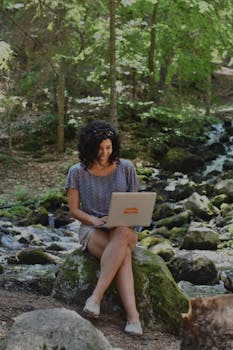 A woman sits working on her laptop by a peaceful forest stream, embodying remote work in nature.