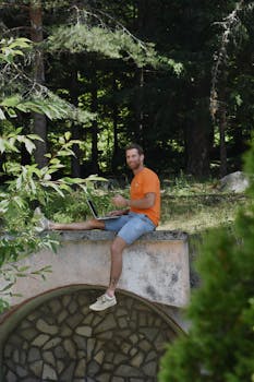 A man in an orange shirt uses a laptop while sitting on a concrete platform in a forest setting.