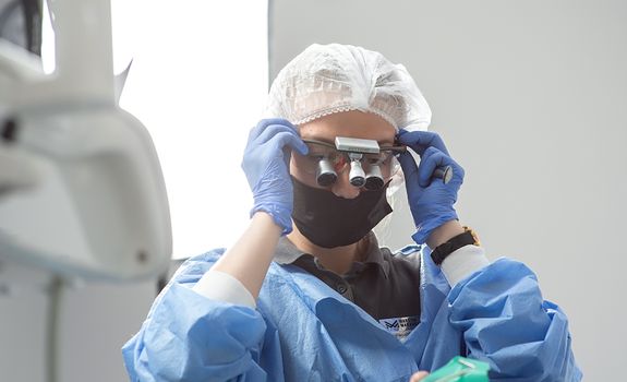 A female dentist adjusts her loupes in a well-lit dental clinic, wearing protective gear for a procedure.