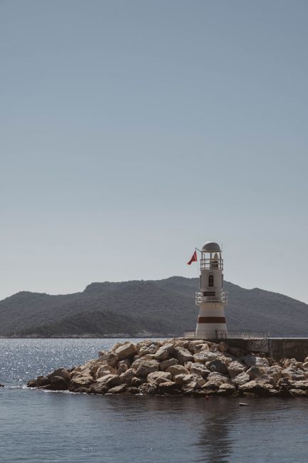 Lighthouse on rocky shore of Kaş, Turkey with serene sea and mountains in the background.