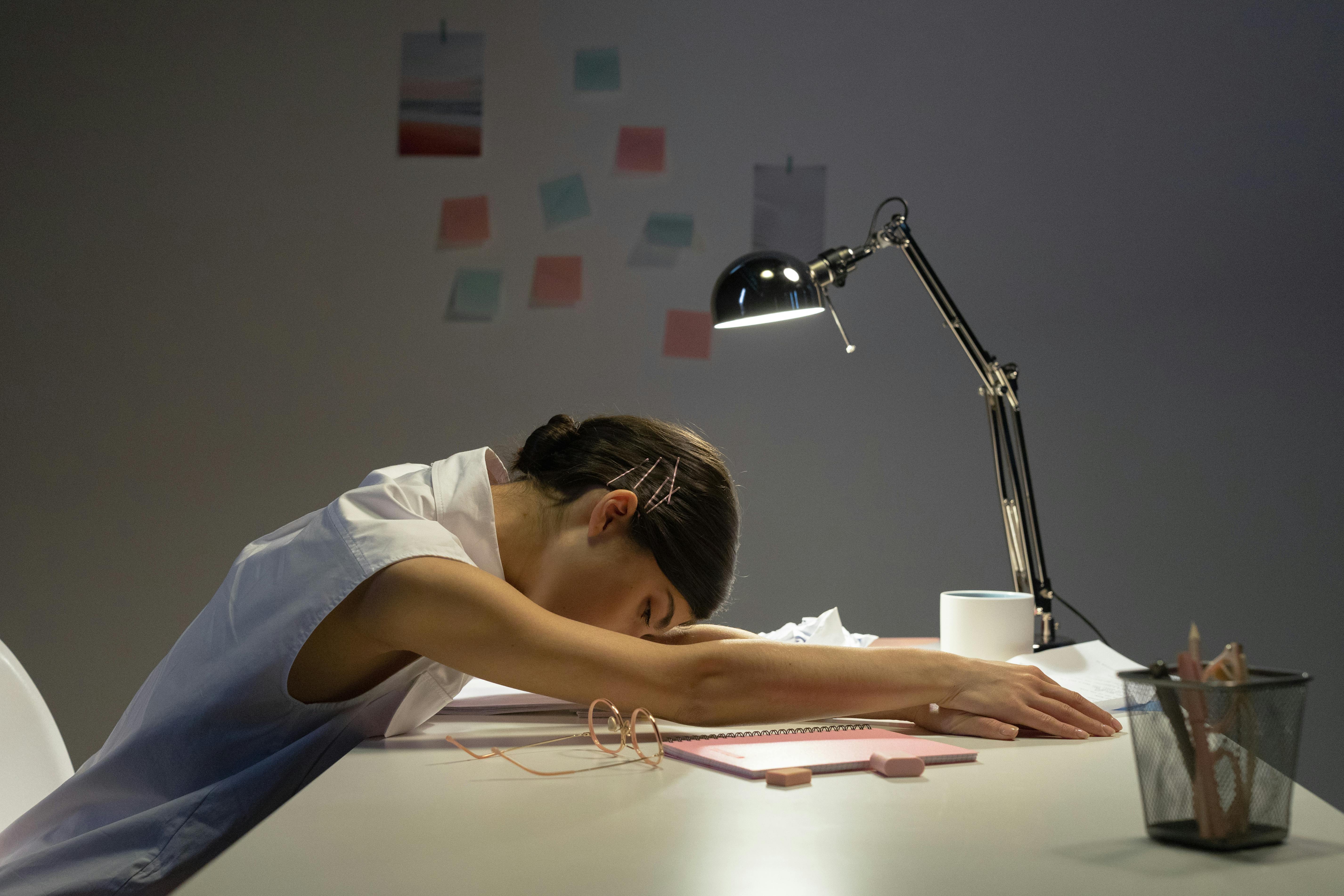 A tired woman rests her head on an office desk under a lamp, symbolizing workplace fatigue.