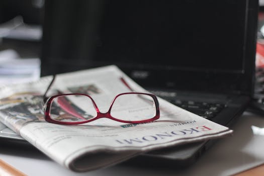 Close-up of red eyeglasses on a newspaper with a laptop in the background, suggesting reading or work.