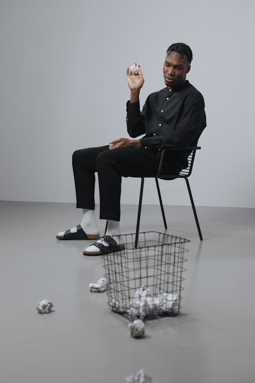 Man seated on a Chair throwing Crumpled Papers on a Trash Bin 