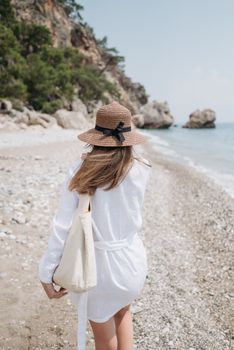Back view of a woman walking along a pebble beach in summer wearing a brown hat and white dress.