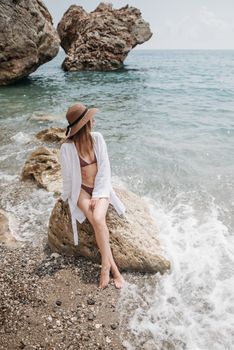 A woman in a swimsuit and hat sitting on a rock by crashing waves, enjoying the beach view.