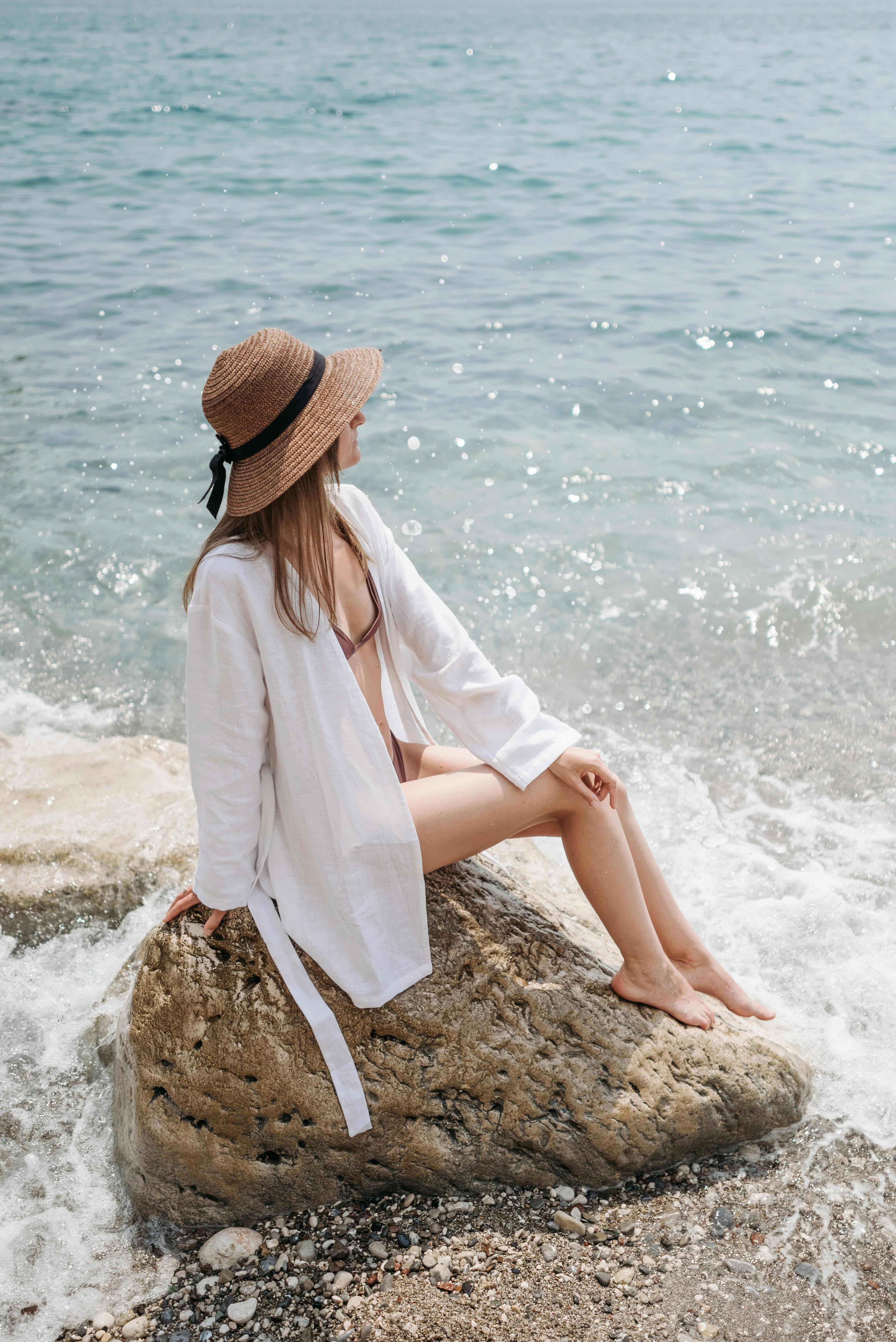 一个穿着泳衣和帽子的女人坐在海边的岩石上，享受夏日