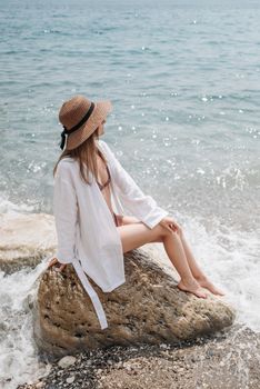 A woman in a swimsuit and hat sitting on a rock by the sea shore, enjoying a summer day.