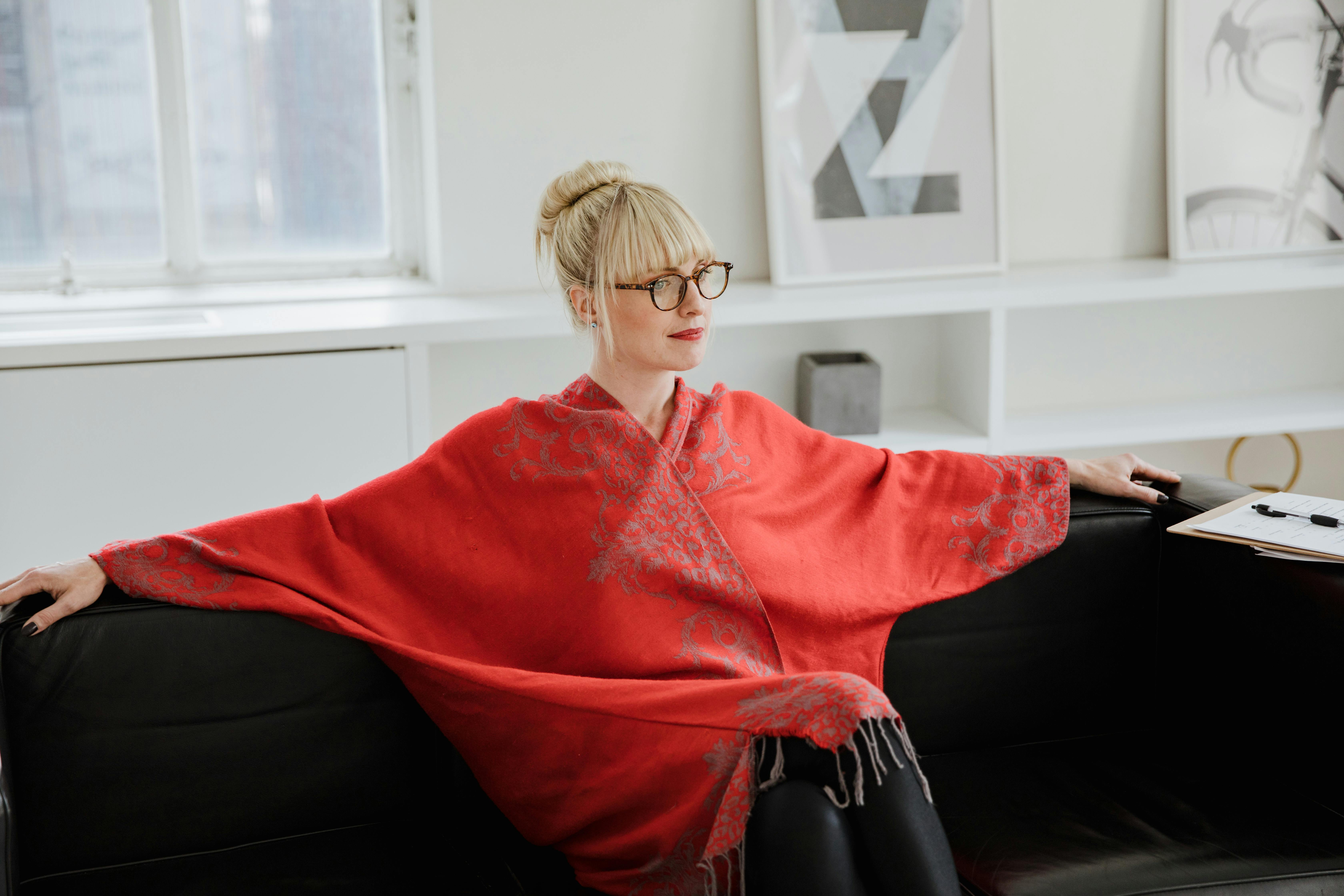 Woman with glasses and blonde hair sits relaxed on a black sofa in a bright room.