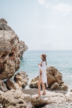 Woman in swimwear and straw hat standing on rocky shore by a calm blue sea.