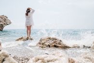Woman in White Dress Shirt and White Skirt Standing on Brown Rock by the Sea during